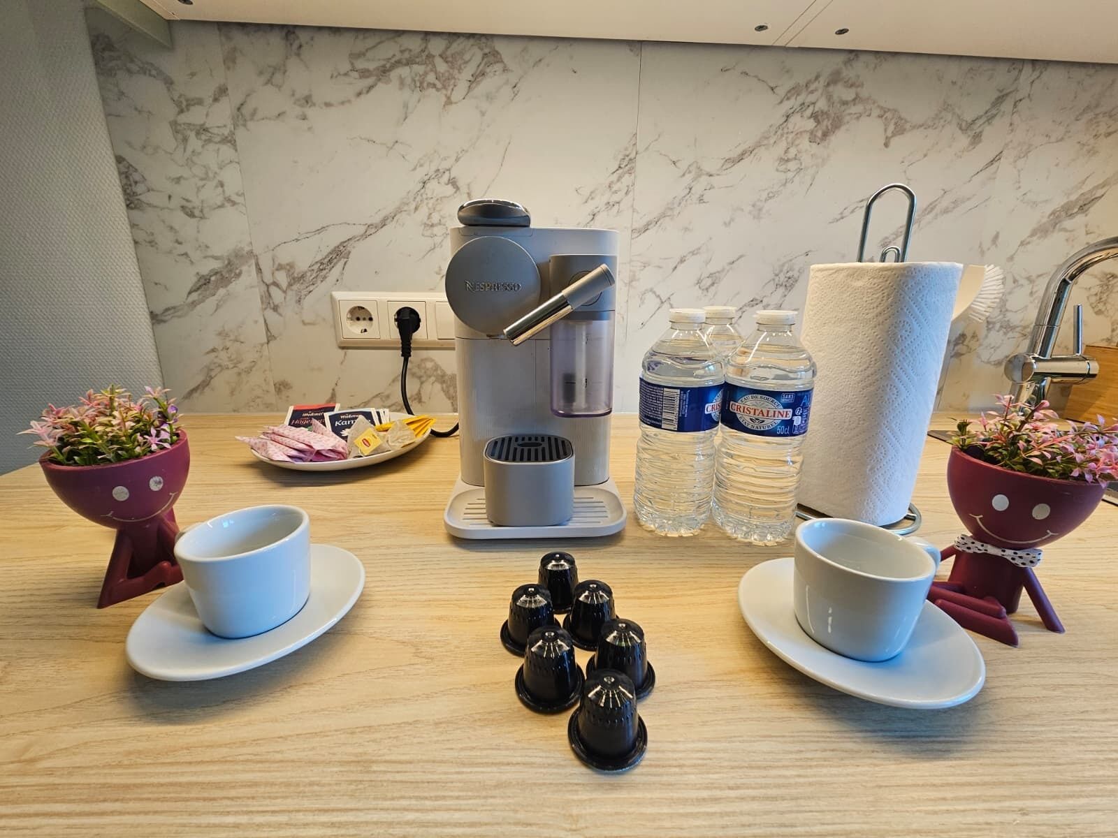 A kitchen counter with a coffee maker, two cups, two bottles of water, a plate with food, and two flower pots.