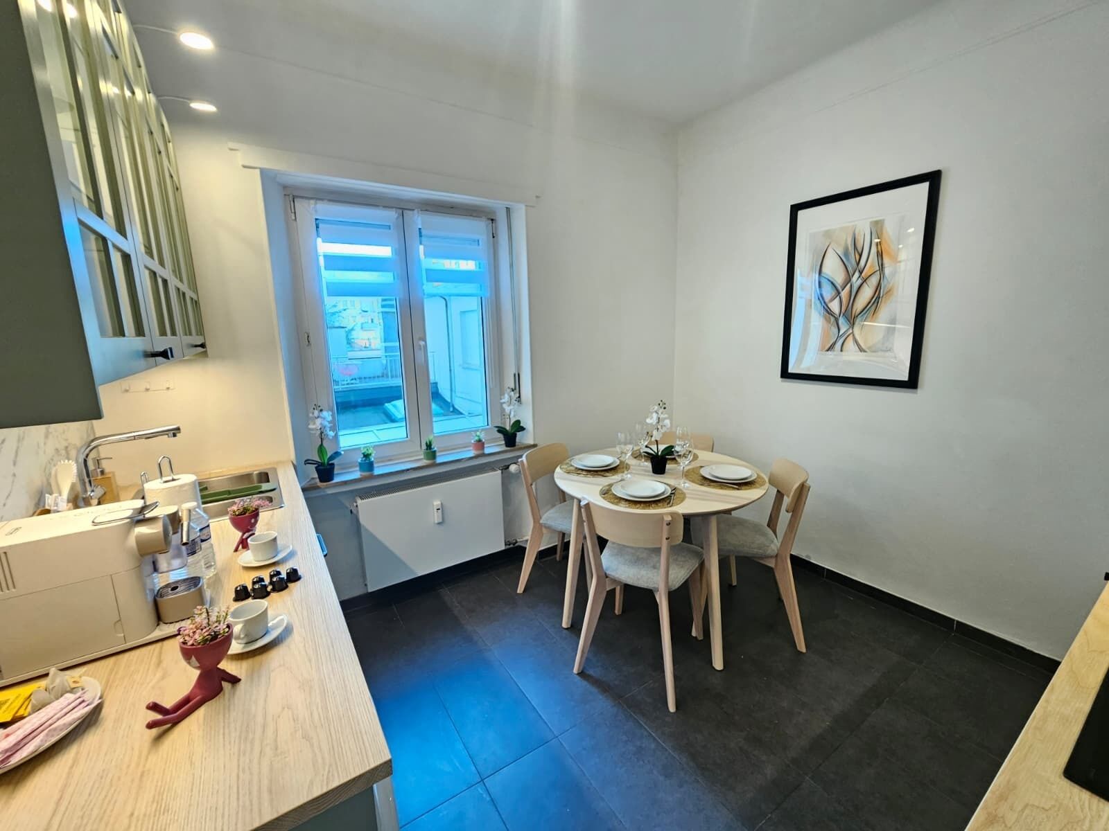 A modern kitchen with a dining area, featuring a window with blinds, a sink, and a countertop with various items.
