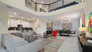 A spacious living area with a white sofa, a coffee table, and a red chair. There is a kitchen in the background with white cabinets and a dining area with a table and chairs. - Disney Villa w/ Pool & 2 Resort Clubhouses (Kissimmee)