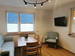 Ein Wohnzimmer mit Esstisch, zwei Stühlen, einer Couch, einem an der Wand befestigten Fernseher und einem Fenster mit Jalousien.