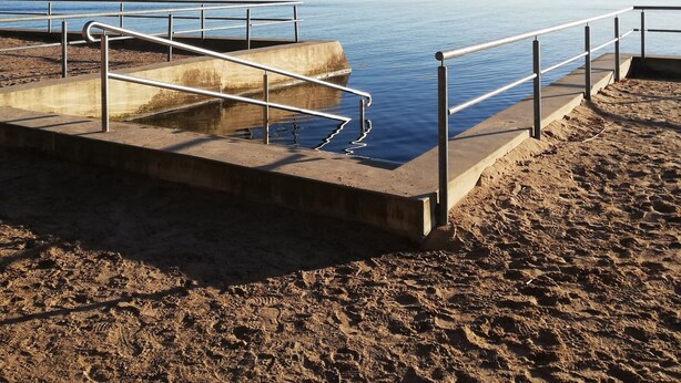 Un chemin piétonnier en béton, bordé de rampes métalliques, mène à une étendue d’eau paisible.