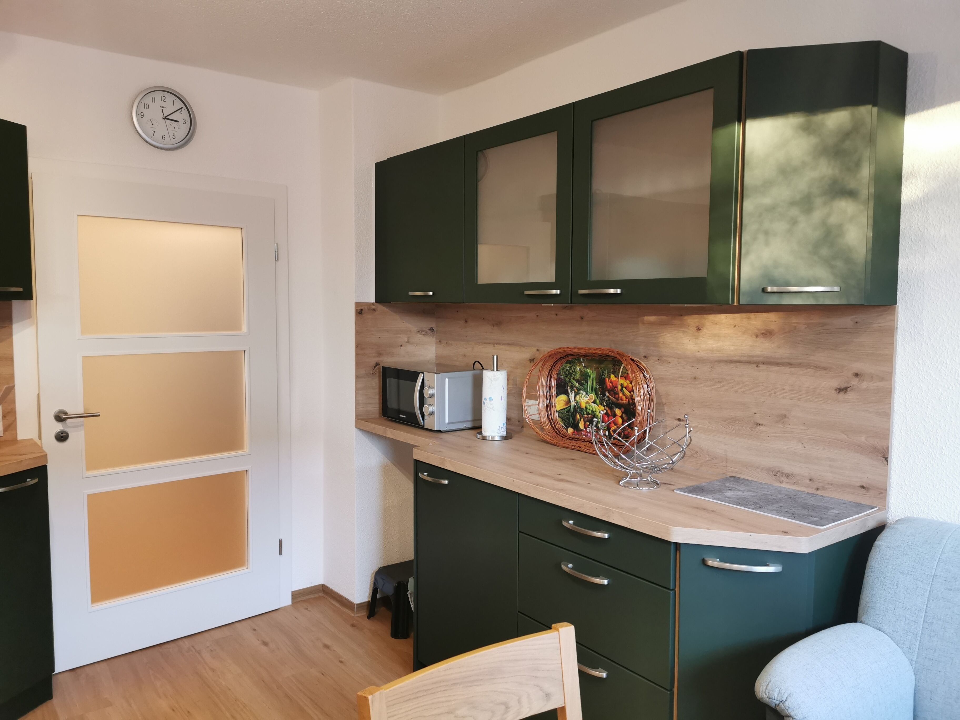 Een kleine keuken met donkergroene kastjes, een magnetron, een rieten mand met groenten en een klok aan de muur.