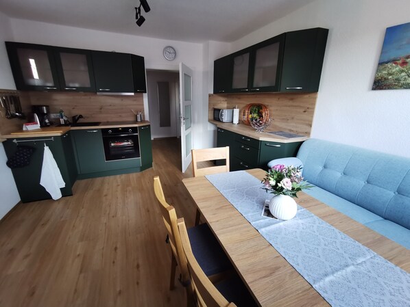 Eine moderne Küche mit dunkelgrünen Schränken, einer Holzarbeitsplatte und einem eingebauten Backofen. Es gibt einen Essbereich mit einem Holztisch, Stühlen und einem hellblauen Sofa.