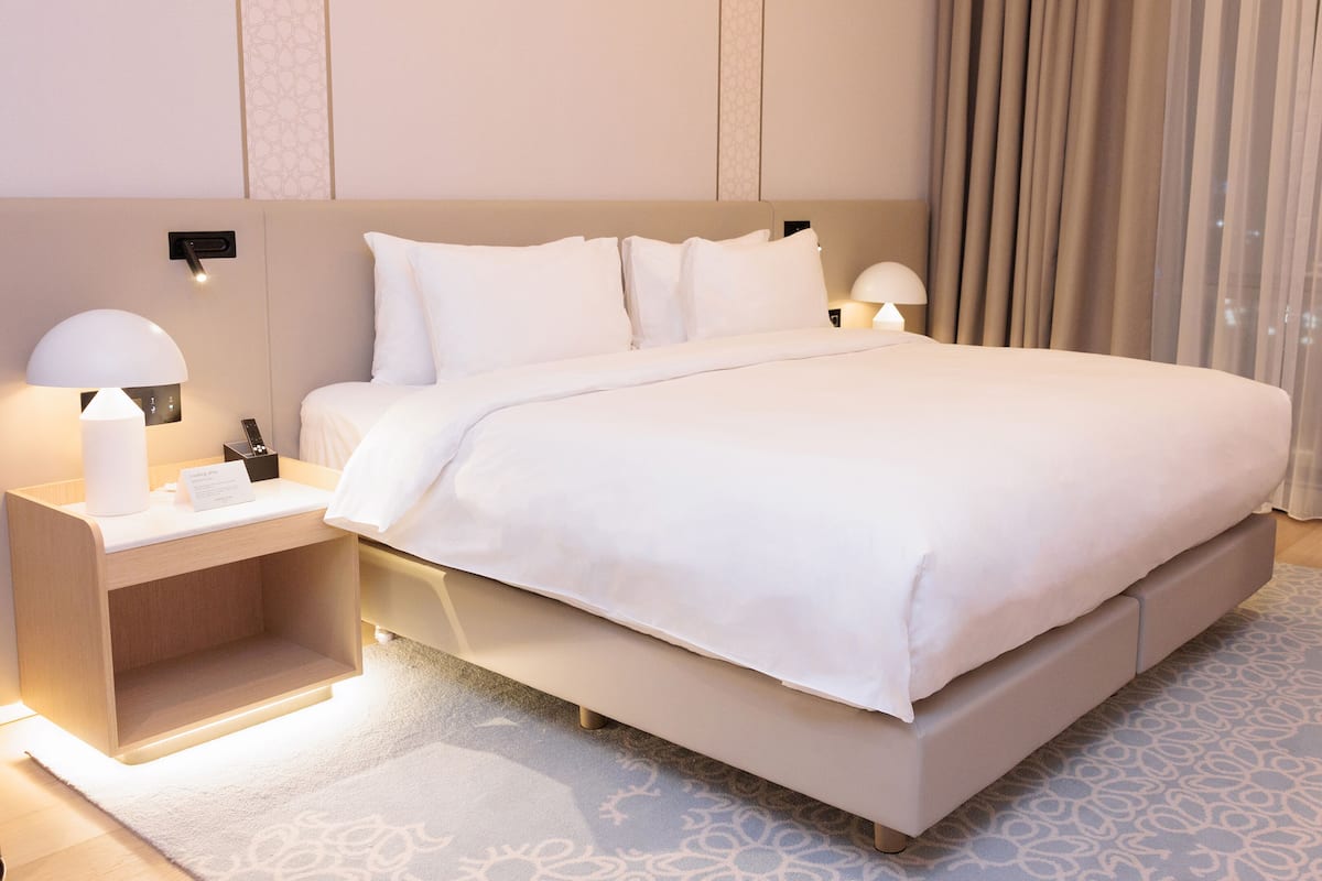 A modern hotel room with a large bed, bedside tables with lamps, a chair, and a view of the city through the window.