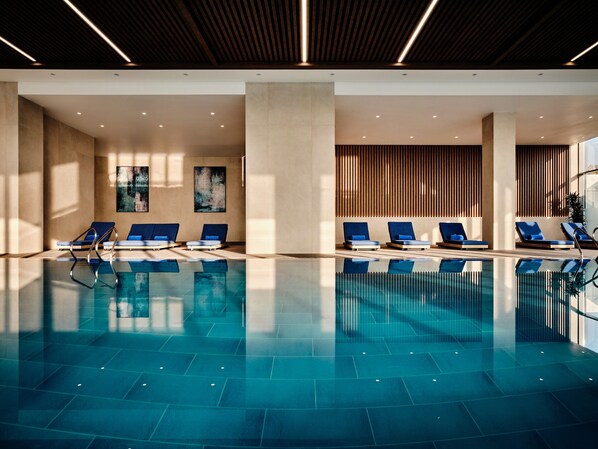 A modern indoor swimming pool with clear blue water, surrounded by lounge chairs with blue cushions, and a wooden slatted wall in the background. - Crowne Plaza Dushanbe by IHG (Dushanbe)