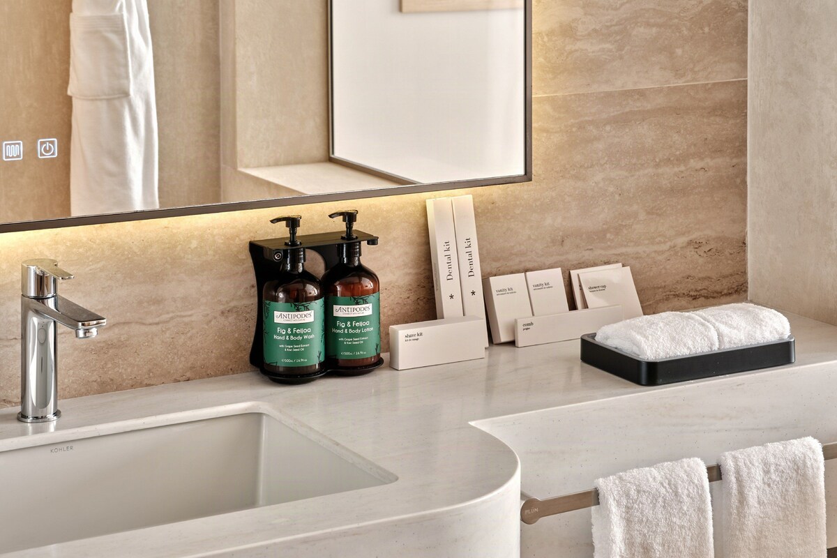 A modern bathroom with a curved sink, a mirror, and a vanity area. There are two hand and body wash bottles, a dental kit, a vanity kit, a shower cap, and a comb.
