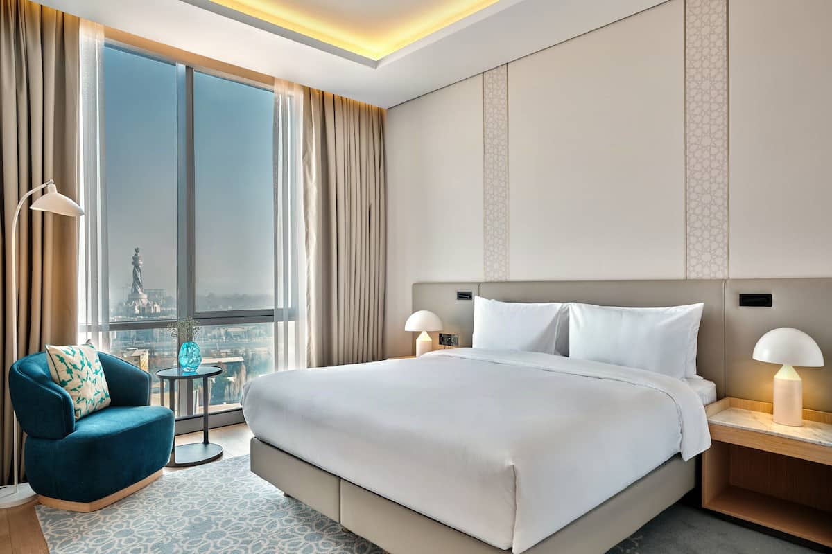 A hotel room with a large bed, two bedside lamps, a chair, a small table, and a view of a cityscape through a large window.