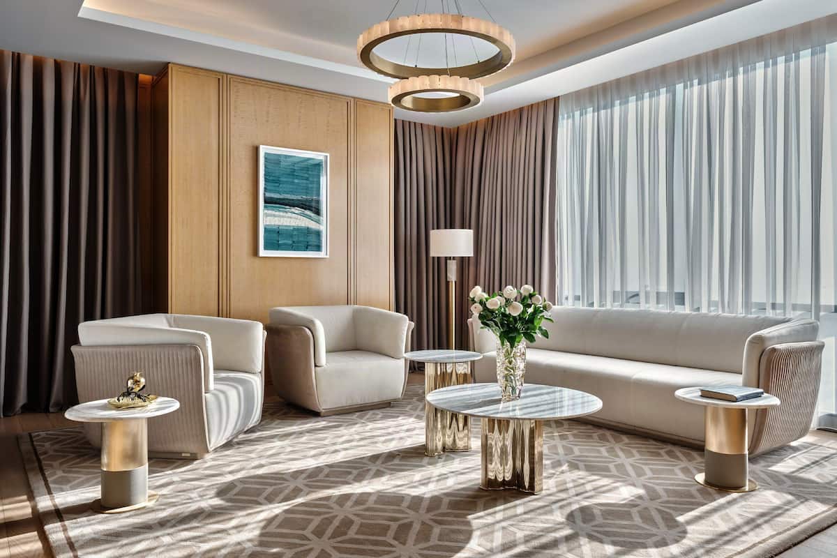 A modern living room with a sofa, two armchairs, a coffee table, and a floor lamp. There is a painting on the wall and a window with curtains.