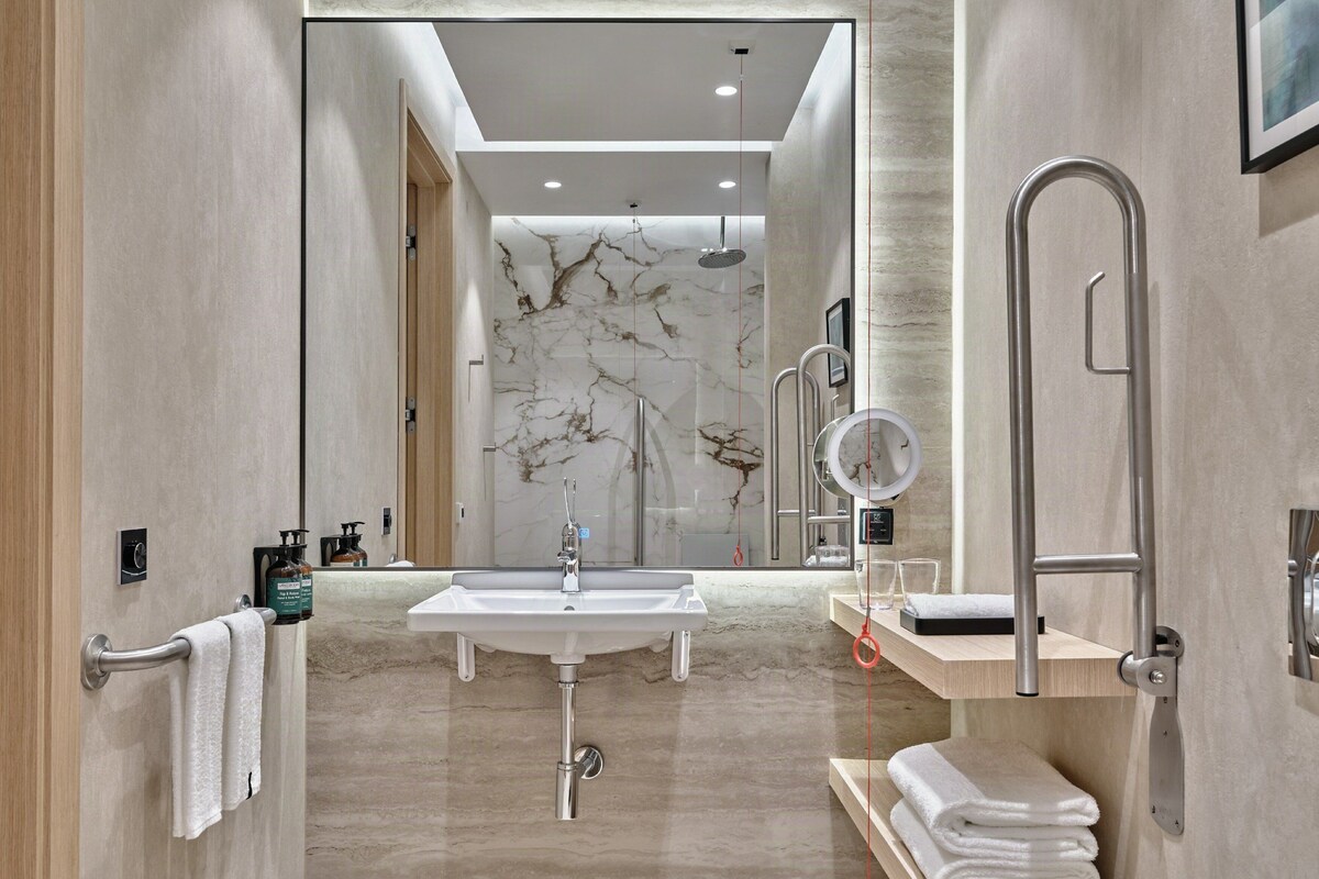 A modern bathroom with a large mirror, a sink, and a shower. There are towels on a shelf and a towel rack on the wall.