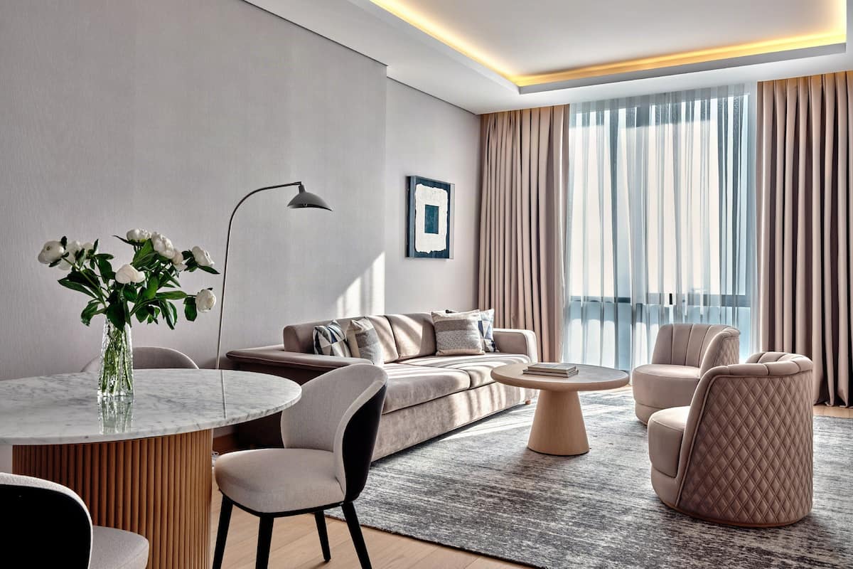 A modern hotel room with a sofa, armchairs, a round coffee table, and a vase of flowers.