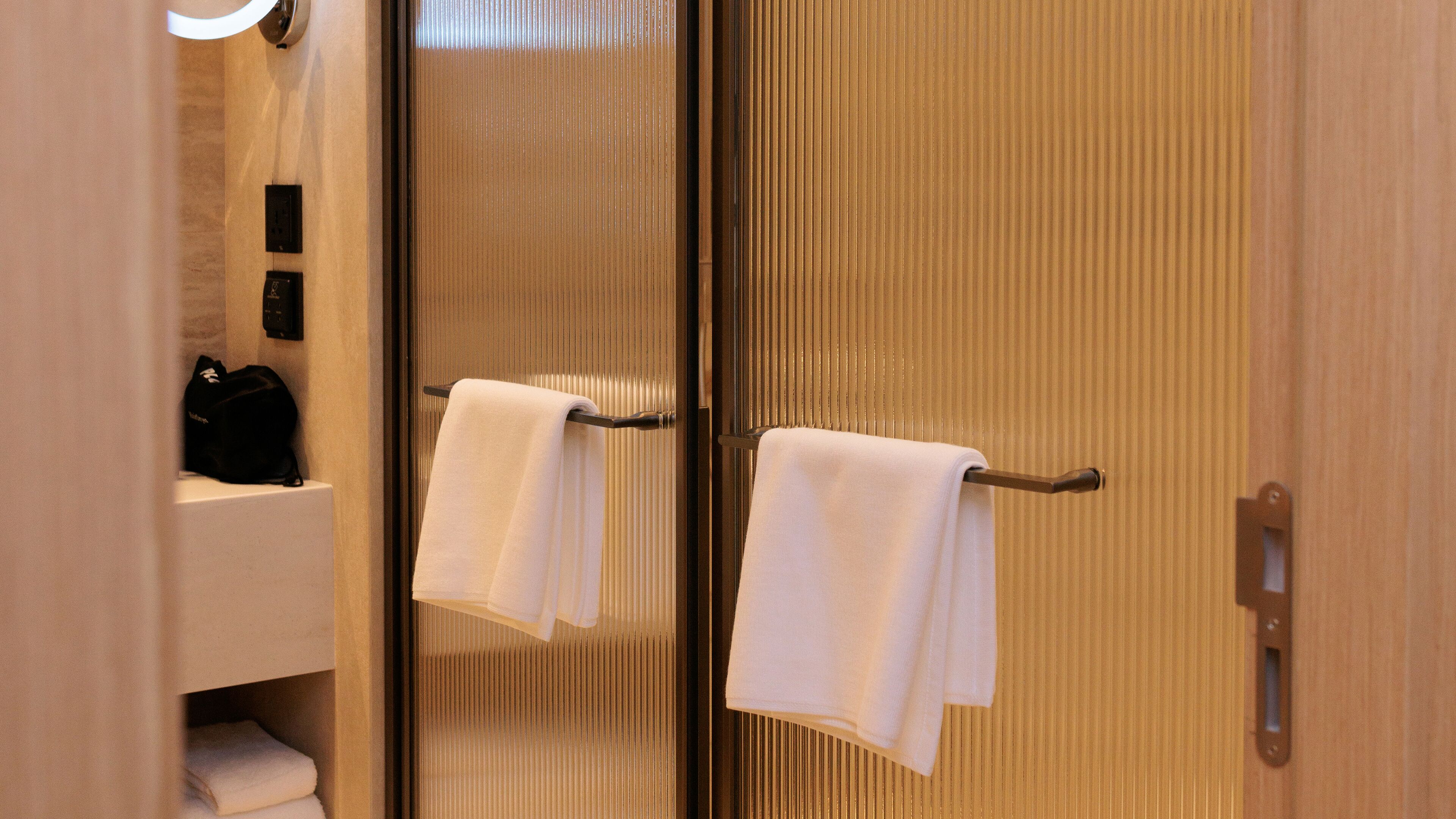 A bathroom with a wooden door, a towel rack with two white towels, and a black bag on a shelf.