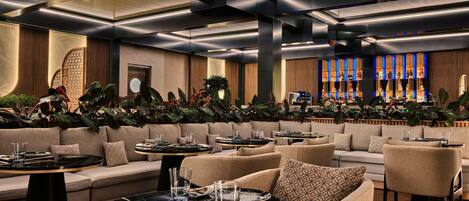 Un área de lounge moderna con sofás, mesas redondas y una barra con estantes iluminados.