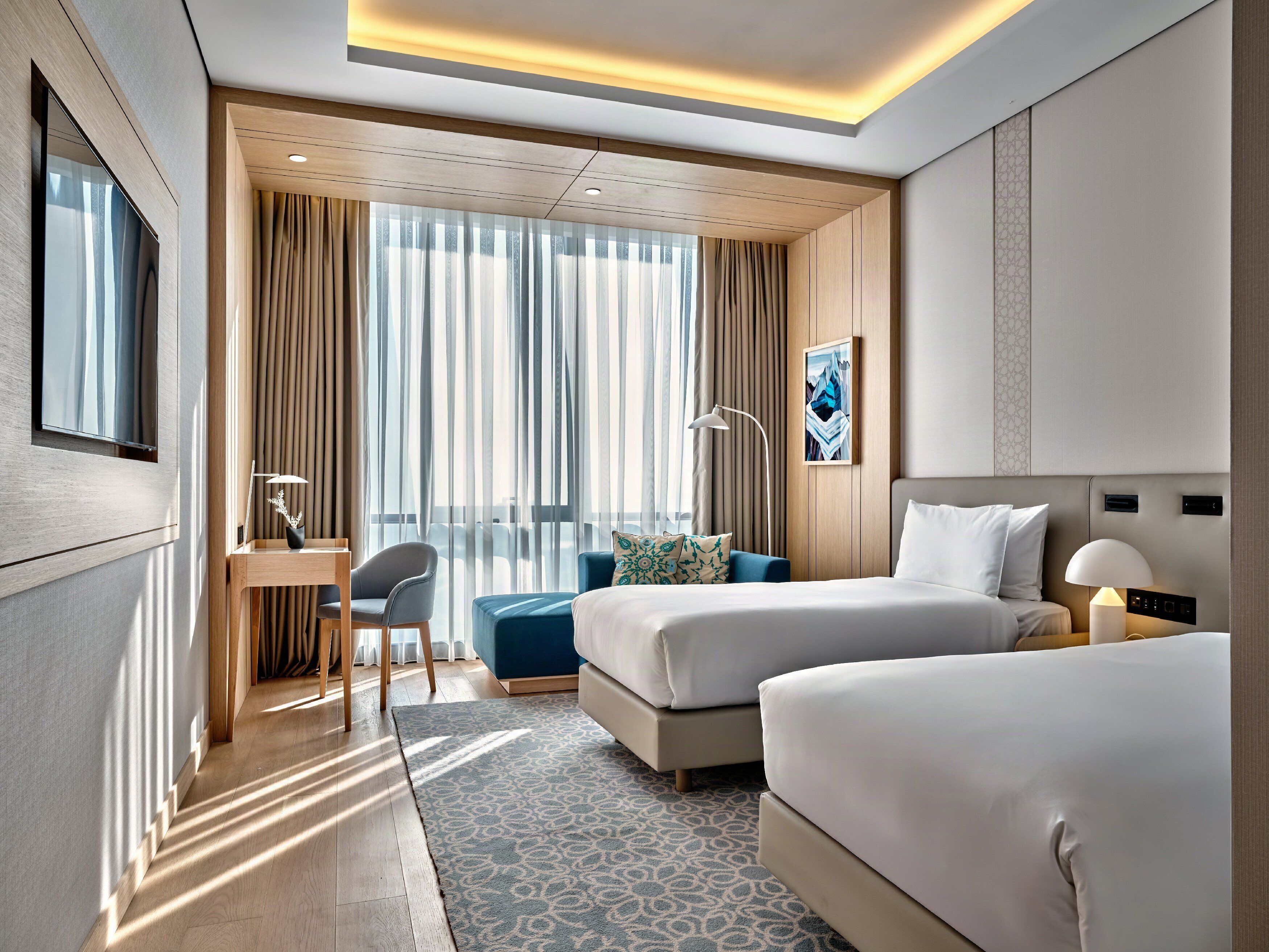 A hotel room with two beds, a desk, a chair, a lamp, and a large window with curtains.