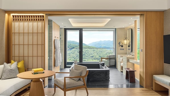 Une chambre d’hôtel moderne dotée d’une grande fenêtre offrant une vue sur les montagnes, d’un coin salon avec une table et une chaise, d’un canapé et d’une salle de bain séparée avec un lavabo et un miroir.