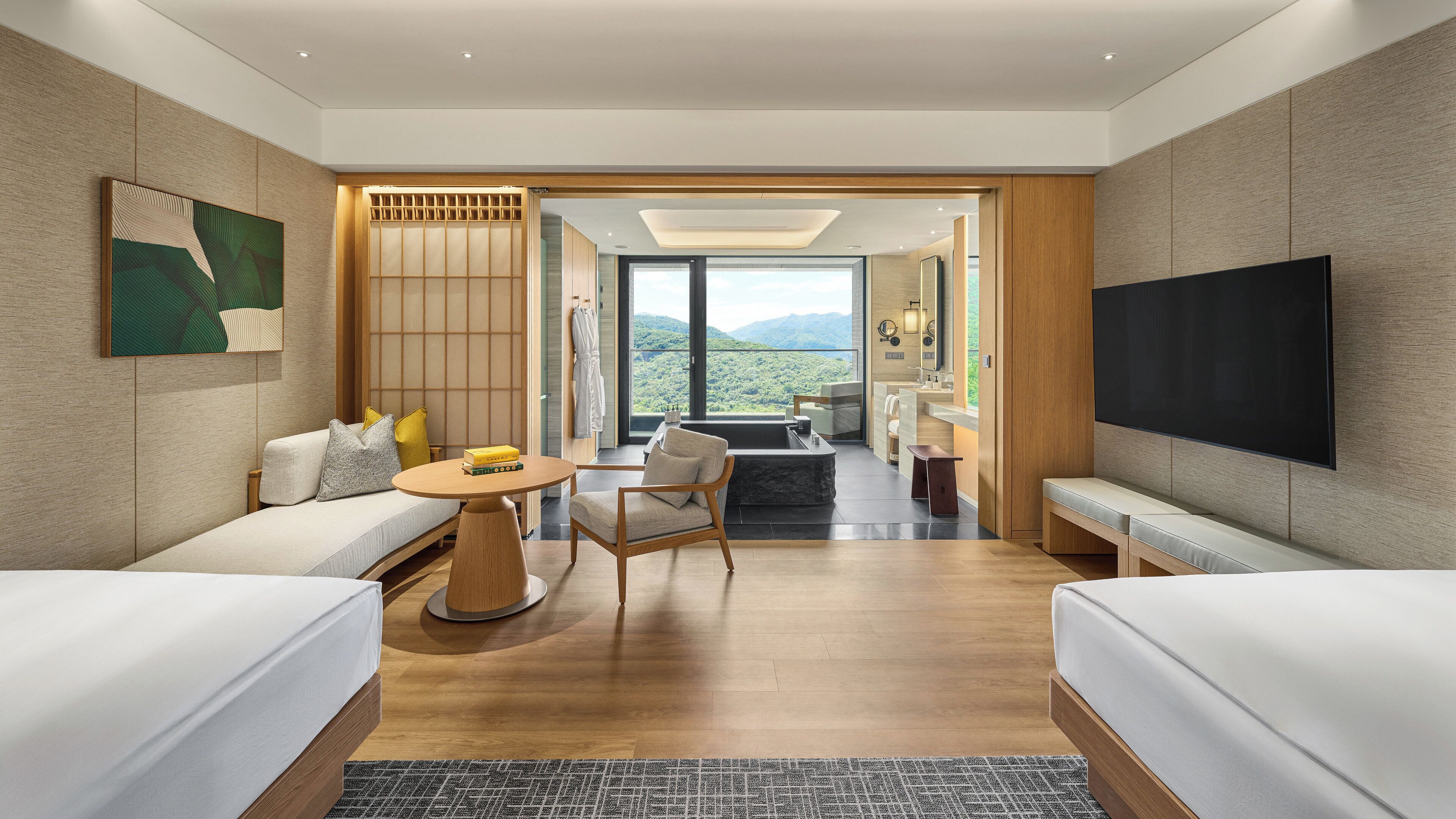 Quarto de hotel moderno com sofá, uma pequena mesa redonda, uma cadeira, uma televisão de tela plana grande e vista para as montanhas pela janela.