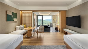 Habitación de hotel moderna con sofá, una pequeña mesa redonda, una silla, un televisor de pantalla plana grande y vistas a la montaña desde la ventana.