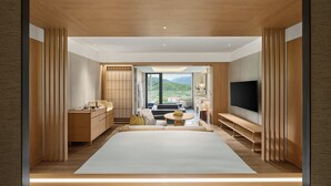 Una habitación de hotel moderna con una cama grande, un televisor, una zona de estar con sofá y una mesa, y vistas a las montañas por la ventana.