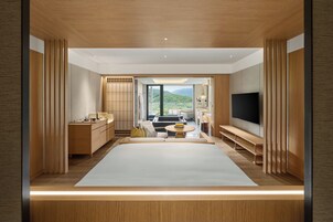 A modern hotel room with a large bed, a TV, a seating area with a sofa and a table, and a view of the mountains through the window.