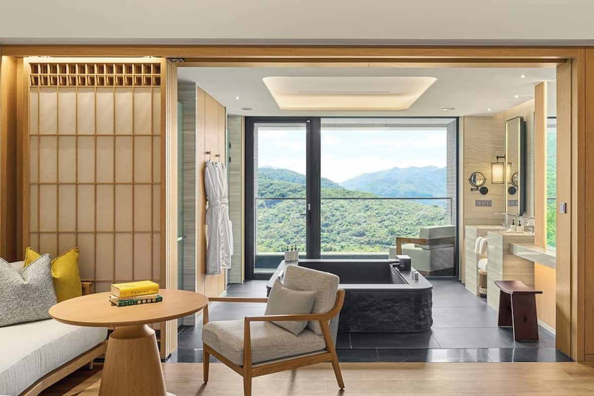 Une chambre d’hôtel moderne dotée d’une grande fenêtre offrant une vue sur les montagnes, d’un coin salon avec une table et une chaise, d’un canapé et d’une salle de bain séparée avec un lavabo et un miroir.
