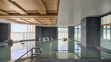 Una piscina interior moderna con paredes de vidrio transparente, vigas de madera en el techo y grandes ventanales que ofrecen vistas al exterior.