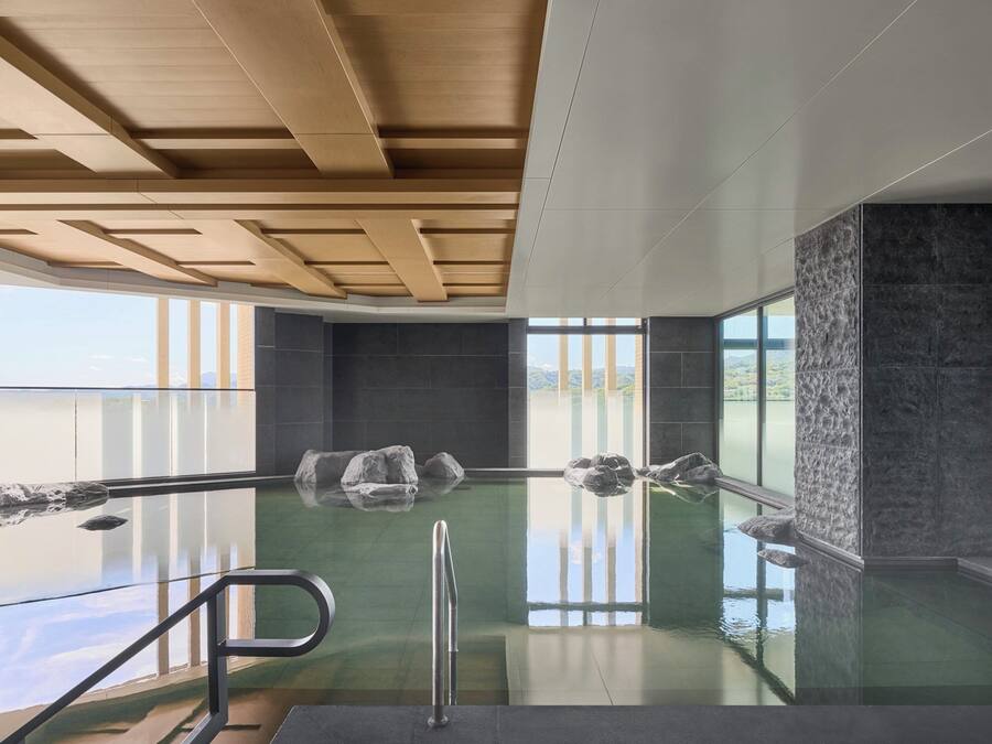Una piscina interior moderna con paredes de vidrio transparente, vigas de madera en el techo y grandes ventanales que ofrecen vistas al exterior.