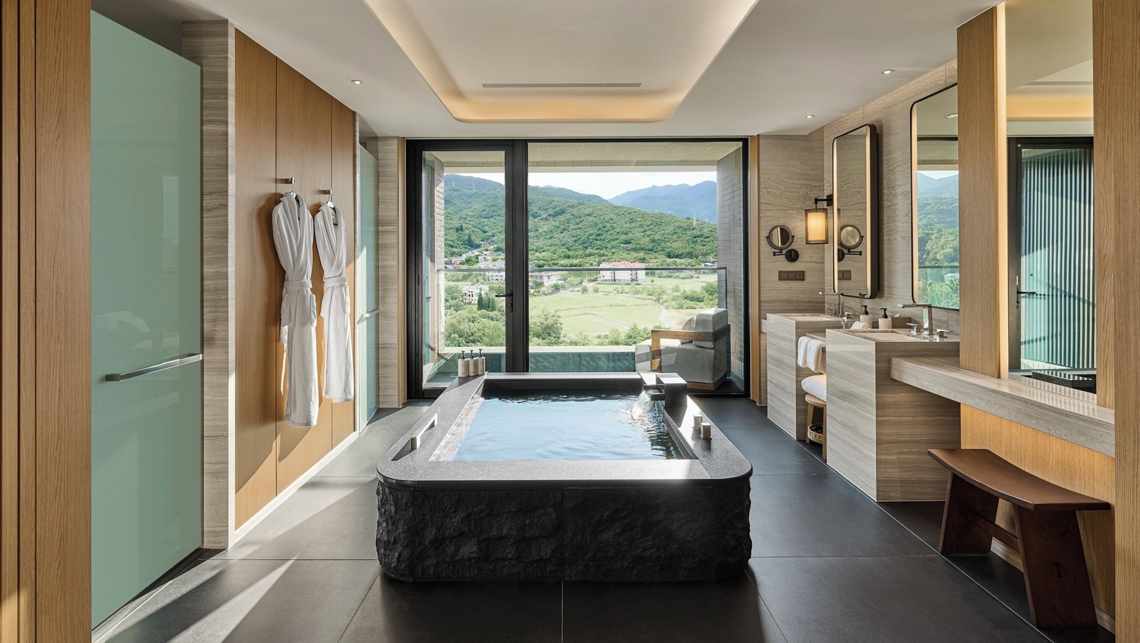 A modern bathroom with a large bathtub, a vanity with a mirror, and a view of a green landscape.