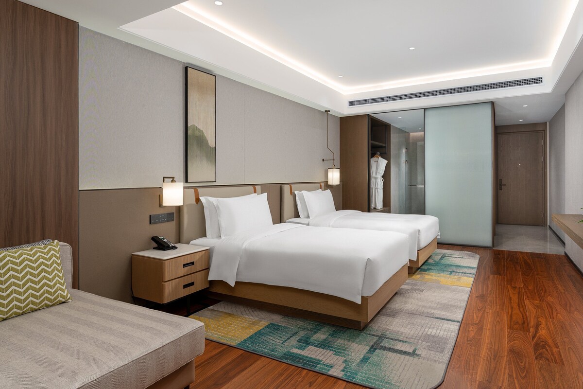 Una habitación de hotel moderna con dos camas, un sofá, una mesita de noche con un teléfono, un espejo y un televisor en la pared.