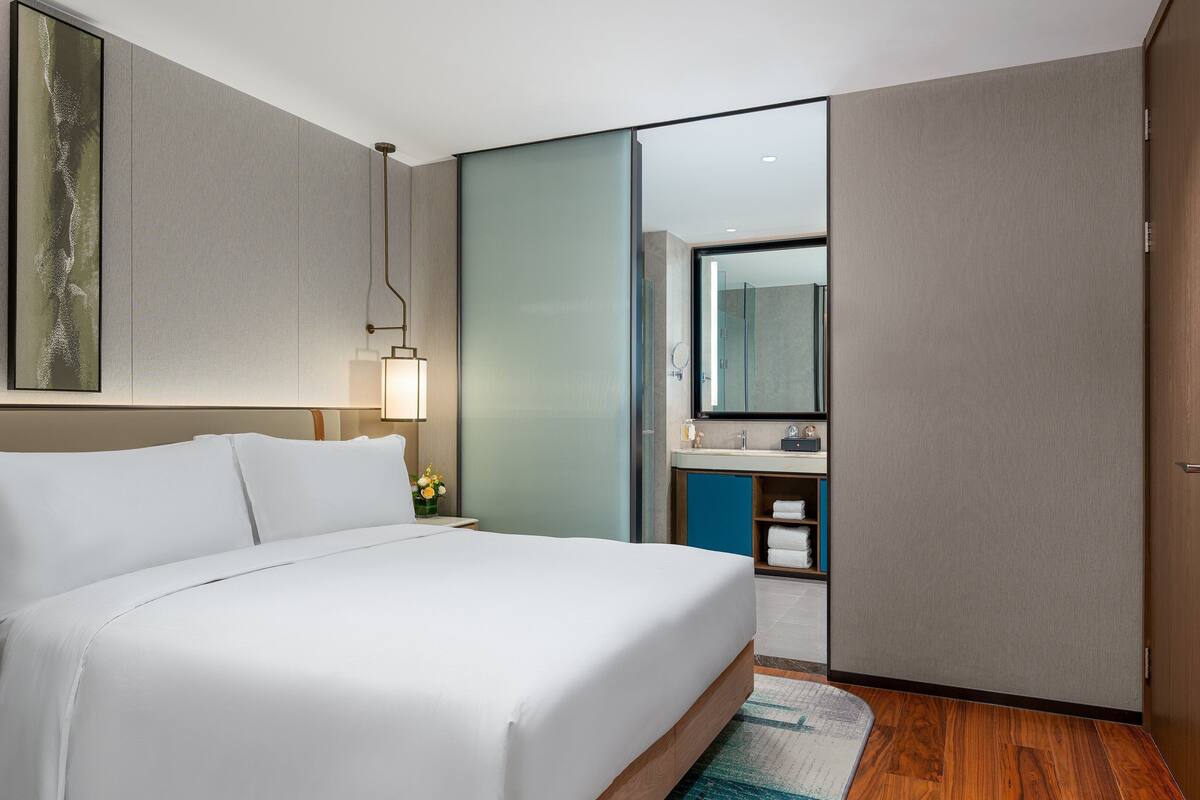Hotelkamer met een groot bed, nachtkastje met een lamp, badkamer met een spiegel en een houten vloer.