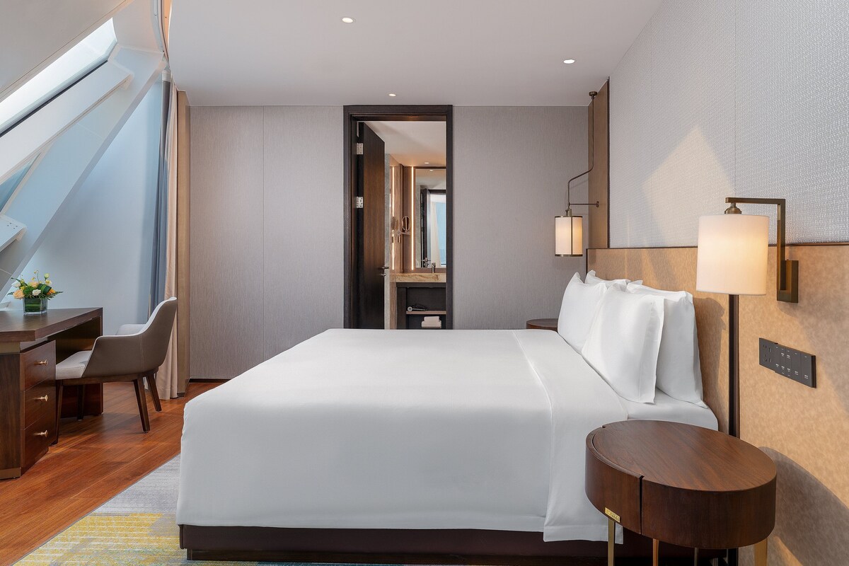 Una habitación de hotel moderna con una cama grande, un escritorio y una silla. Se ve el cielo a través de un tragaluz.