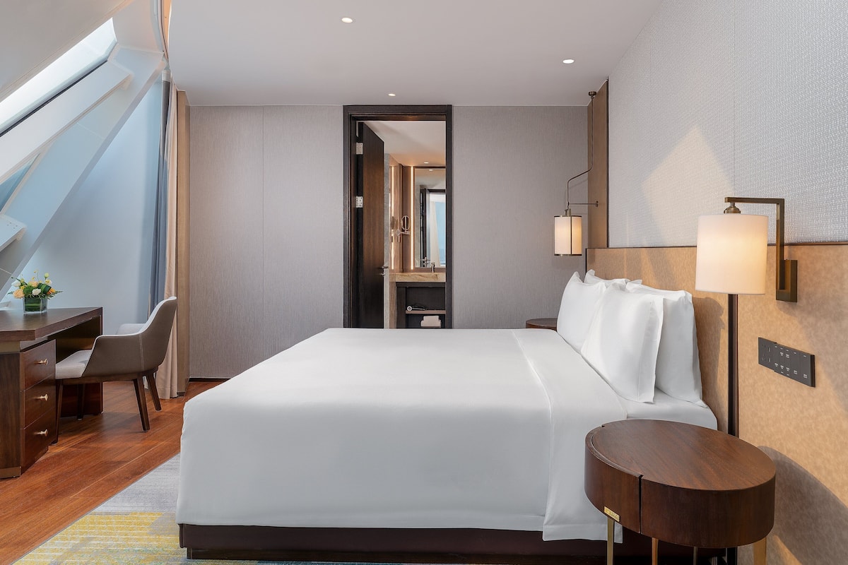 Een moderne hotelkamer met een groot bed, een bureau en een stoel. Er is uitzicht op de lucht door een dakraam.