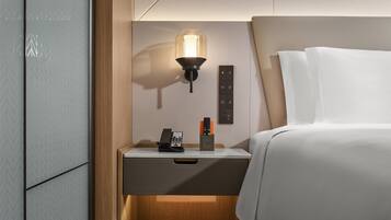 A hotel room with a bedside table, a lamp, a phone, and a control panel.