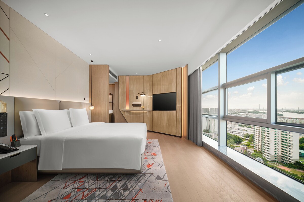 A modern hotel room with a large bed, a TV, a desk, and a view of the city.