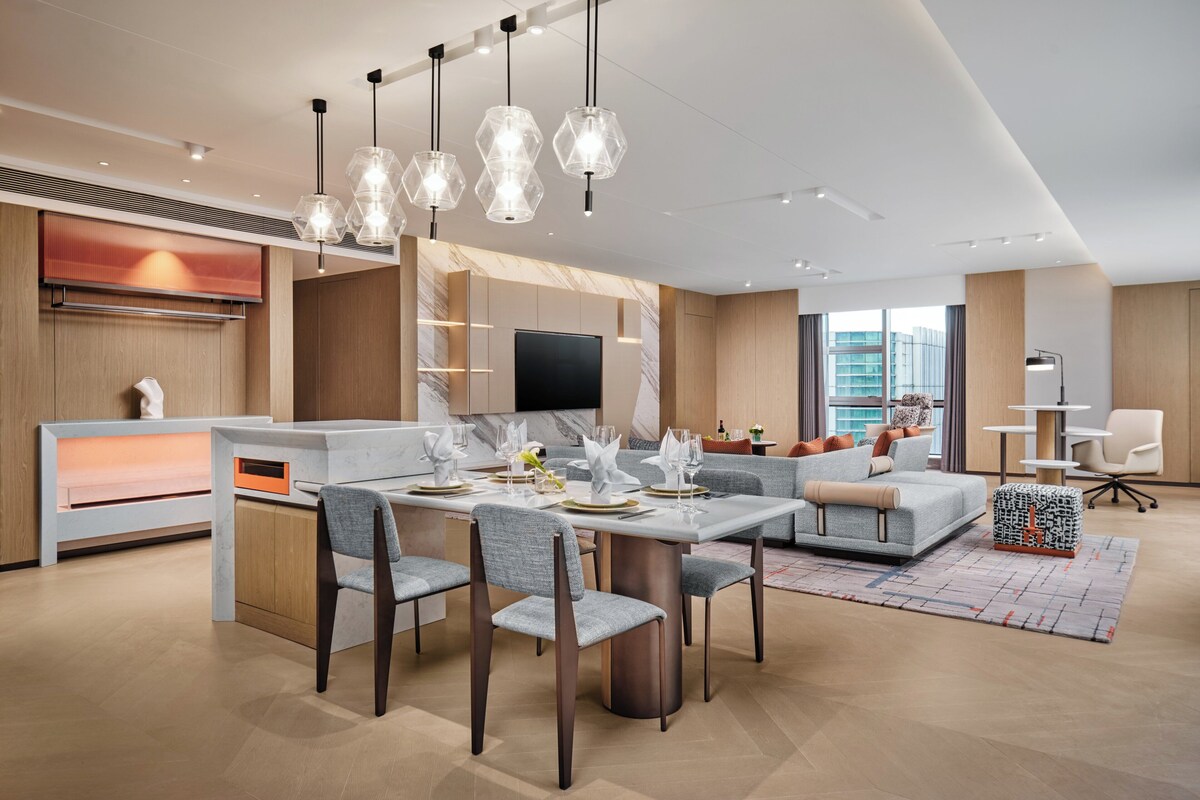 A modern kitchen with a dining area, featuring a marble countertop, a built-in oven, and a flat-screen TV.