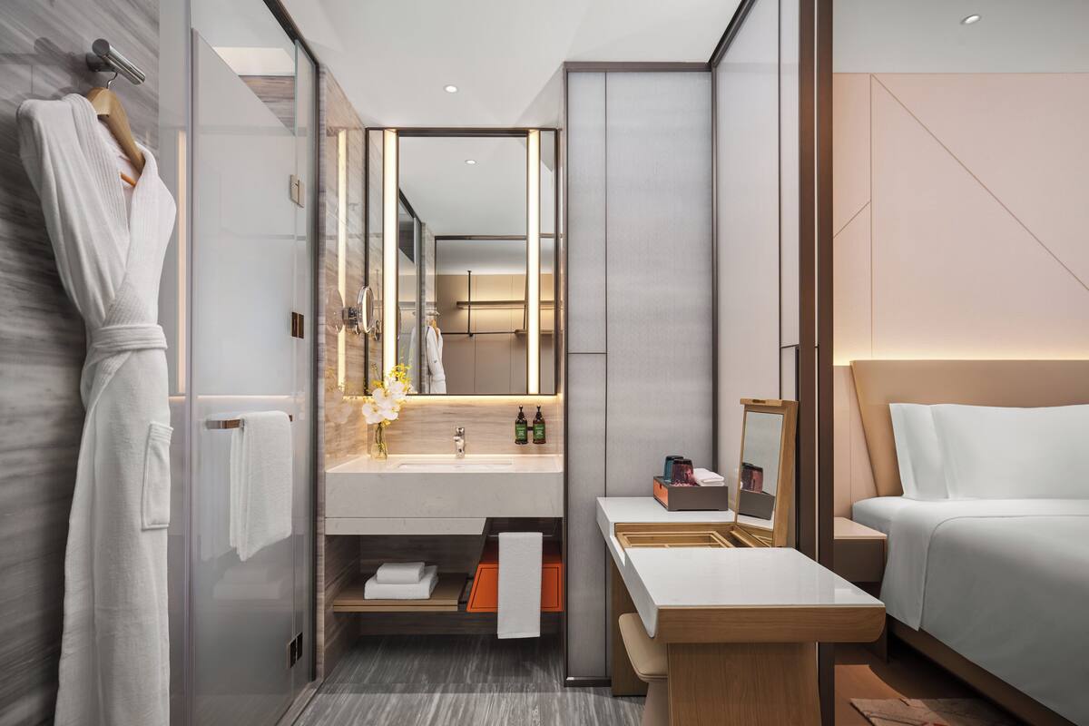 A modern hotel room with a glass shower enclosure, a wooden vanity, a large mirror, a bedside table, and a bed.