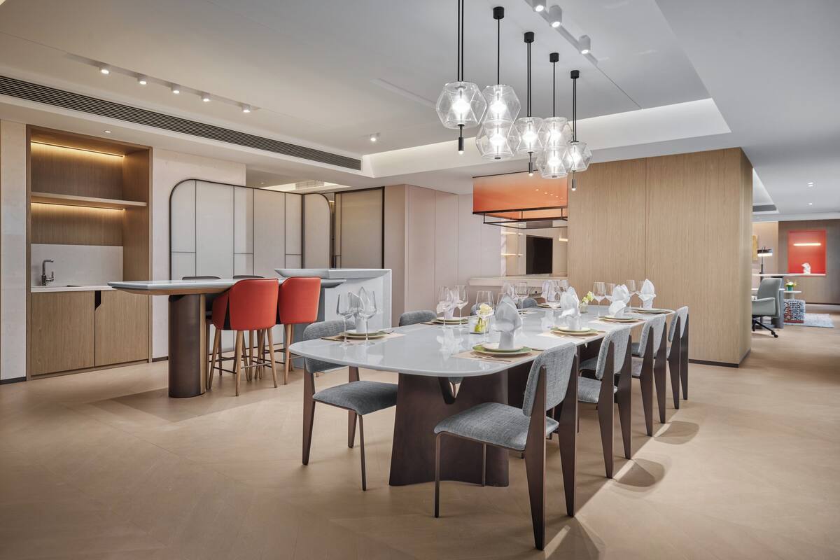 A modern dining area with a white table set for six, surrounded by grey chairs. There are red chairs at a separate table, and a kitchen area is visible in the background.
