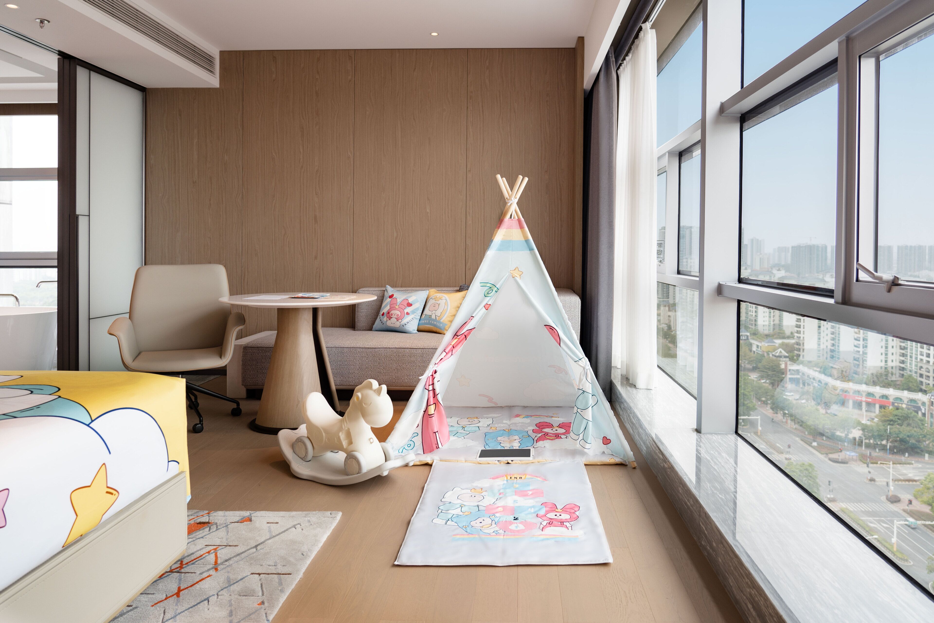 A modern hotel room with a large window, a cozy seating area, a colorful play tent, and a plush toy.