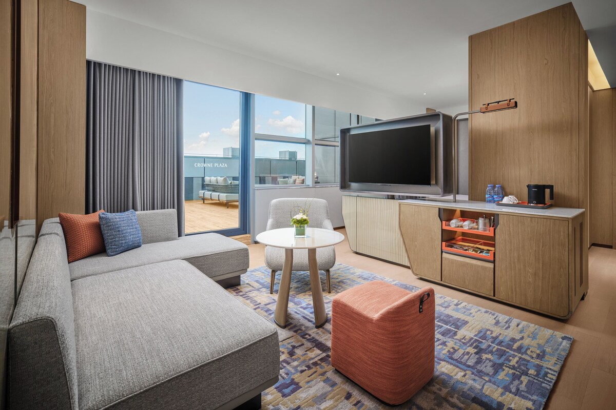 A modern living room with a large sofa, a flat-screen TV, a coffee machine, and a view of the city.