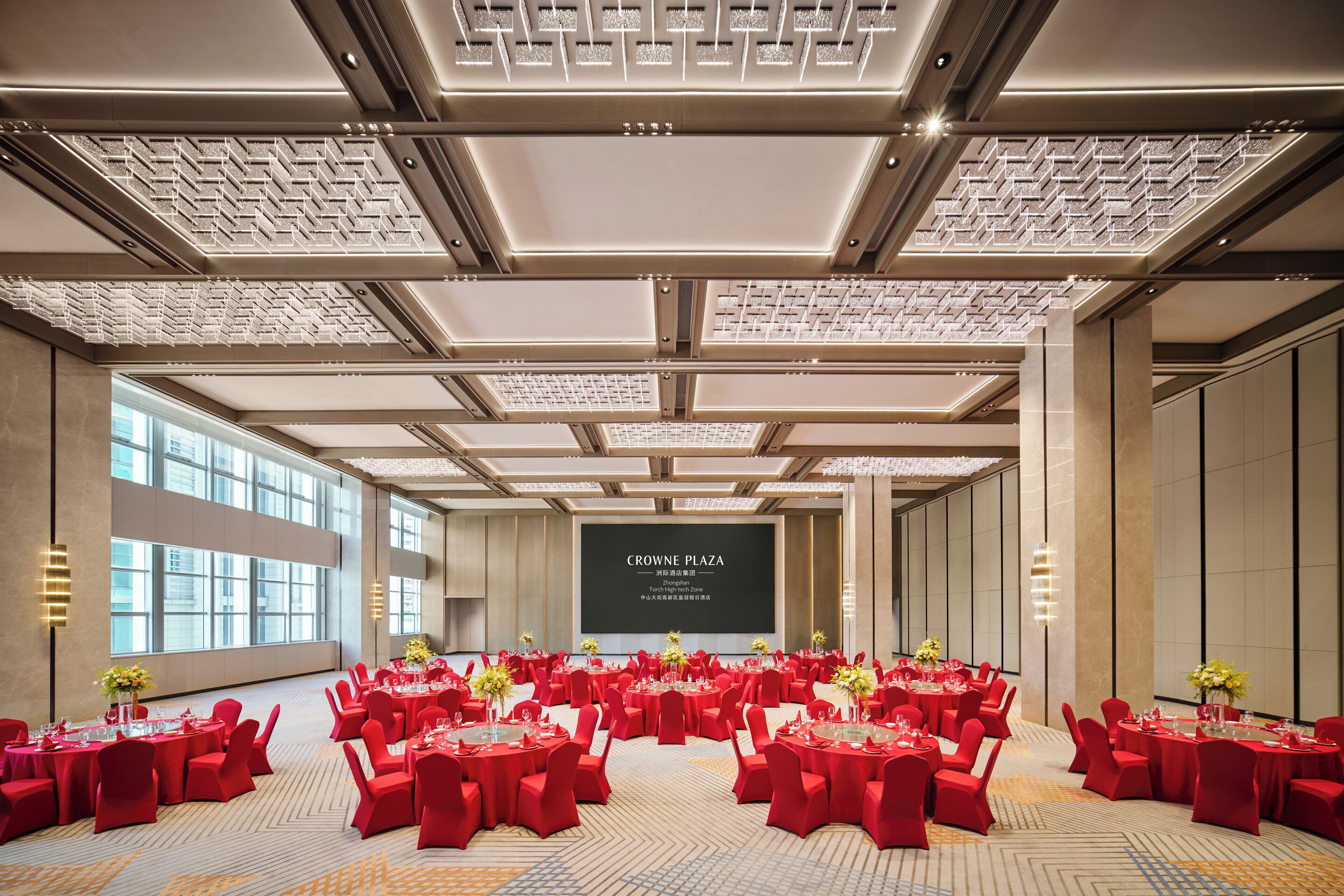 A spacious conference room with multiple round tables, red chairs, and a large screen displaying text.