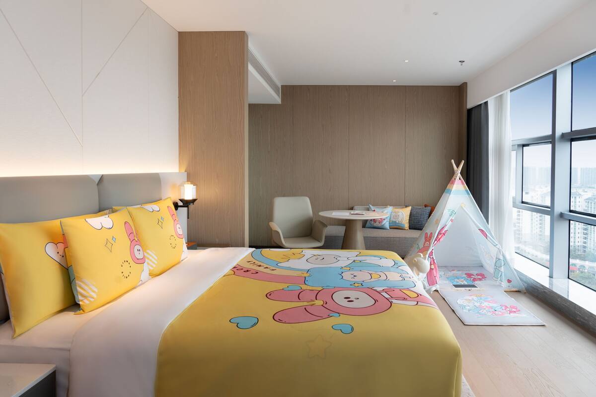 A hotel room with a large bed, a small table, a chair, and a window with a city view.
