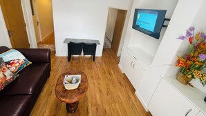 A modern living room with a dark brown leather sofa, a wooden coffee table, a grey console table with two black chairs, a mounted TV displaying a welcome screen, and a white cabinet with a floral arrangement.