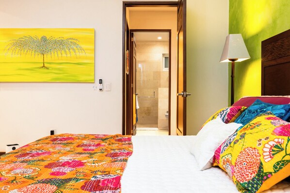 Una habitación de hotel con una colcha de colores, un cuadro de un árbol y un baño visible a través de una puerta abierta.