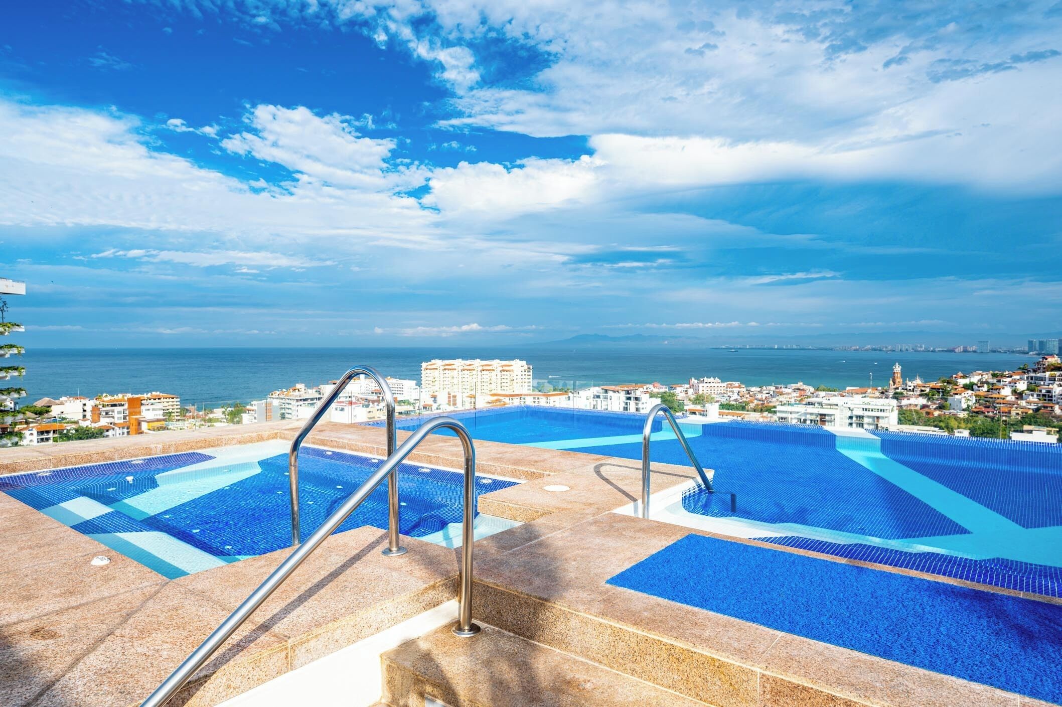 A swimming pool with steps leading down to the water, surrounded by buildings and a view of the sea.
