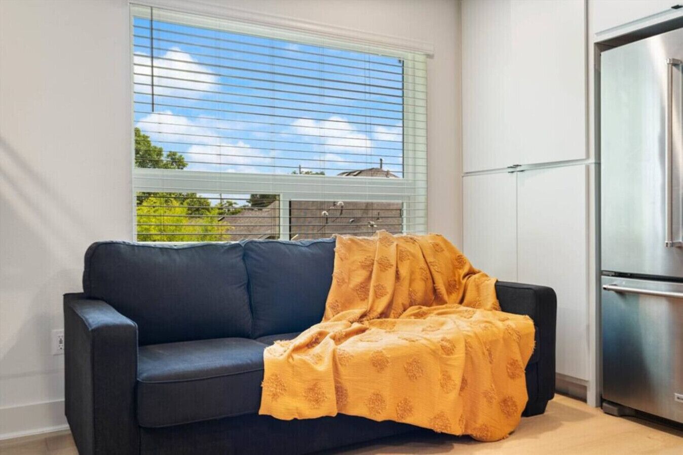 Un salón con un sofá azul oscuro, una manta amarilla estampada, una ventana con persianas y un refrigerador de acero inoxidable.