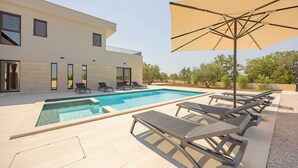 Un área de piscina moderna con piscina rectangular, sillones de descanso y una sombrilla grande.