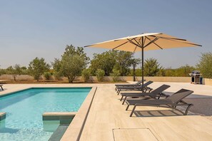 A rectangular swimming pool with clear blue water, surrounded by a light-colored deck. There are several black lounge chairs aligned along the poolside, and a large beige umbrella providing shade.