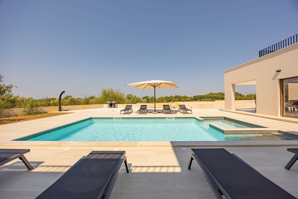 A rectangular swimming pool with a clear blue water surface, surrounded by a light-colored deck. There are four lounge chairs and two umbrellas providing shade. The pool area is adjacent to a modern building with large glass doors.