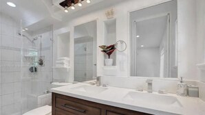 A modern bathroom with a glass shower enclosure, a white toilet, a double sink vanity with dark wood cabinets, and a large mirror above the sink. - LUXTROPICA (Miami)