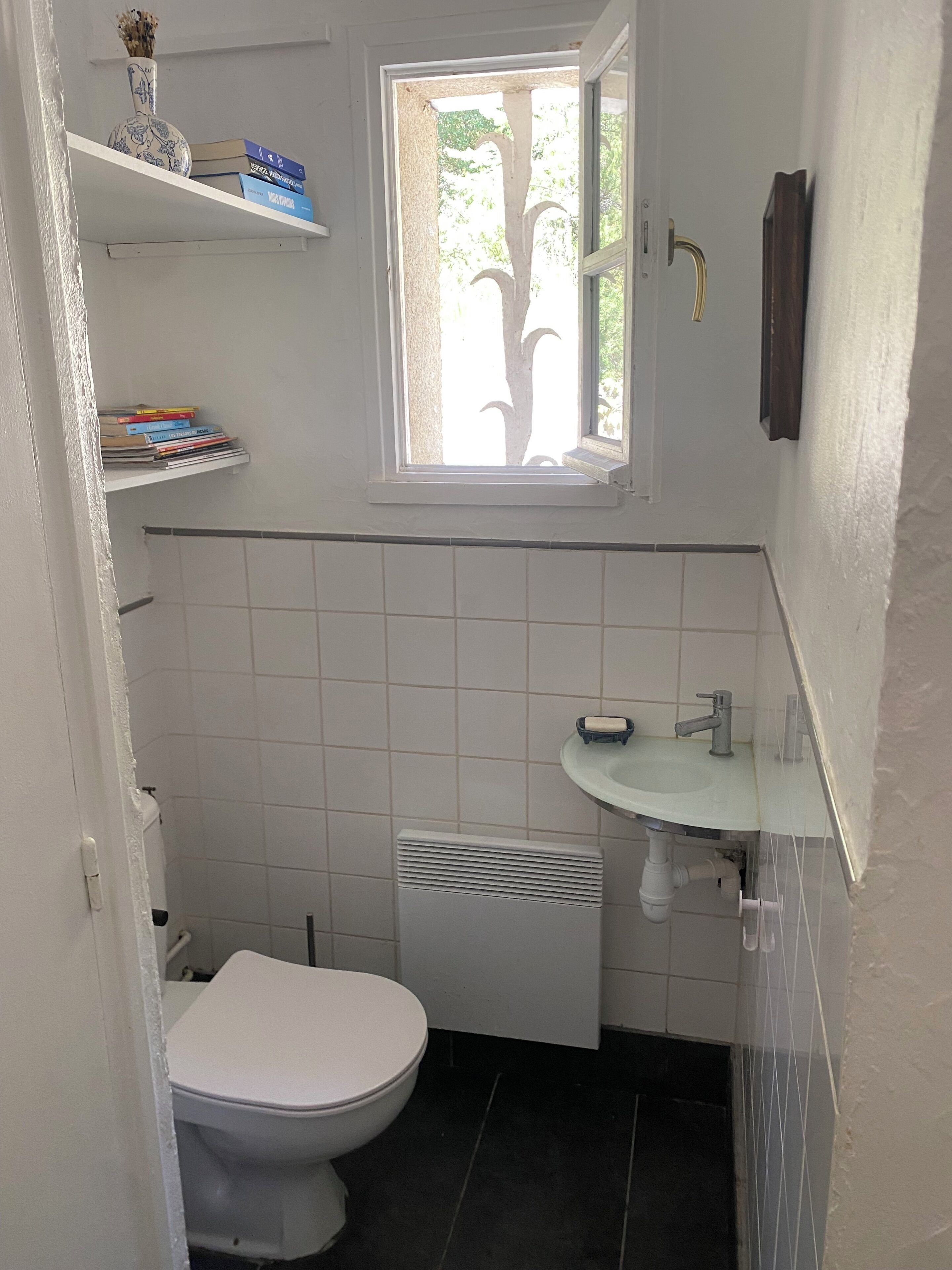 A small bathroom with a toilet, a window, a shelf with books, and a sink.