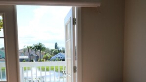 Ein Zimmer mit einer Schiebetür, die auf eine Terrasse mit weißem Zaun und Blick auf Häuser und Grünflächen führt. Dort steht ein beiges Sessel mit dunkler Sitzkissen und ein kleiner Hocker.