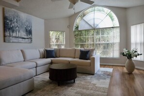 Uma sala de estar com sofá de canto, uma mesa de centro redonda e uma janela grande em arco com persianas.